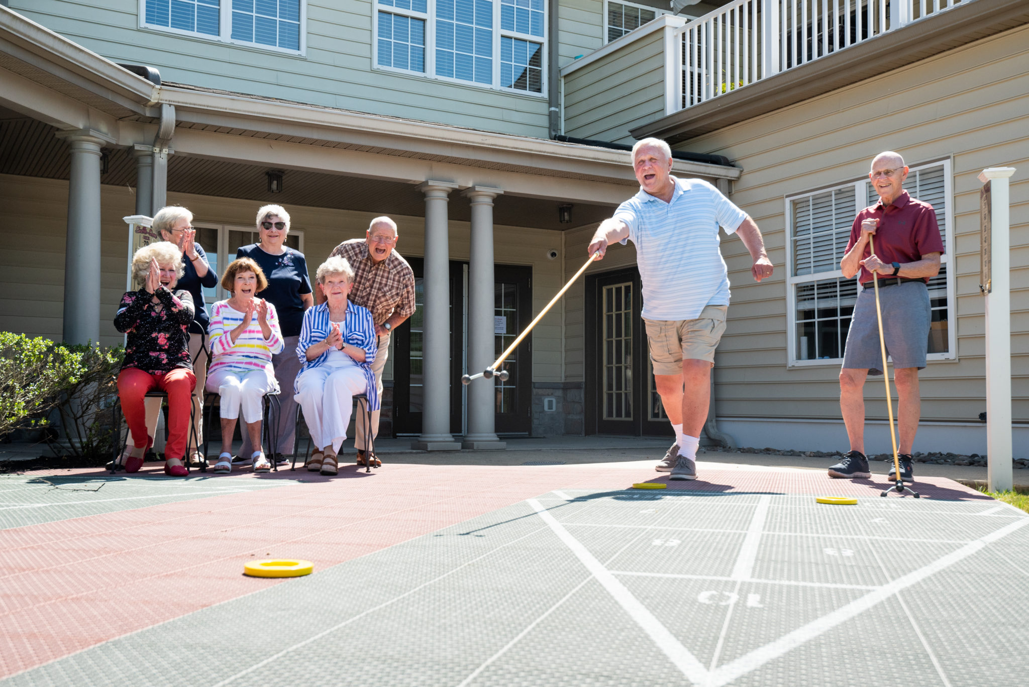 Six residents watch two residents play shuffleboard outside.